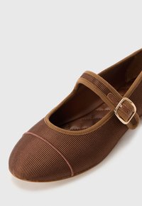 Brown Mary Jane shoe with ribbed texture, round toe, and a strap with a metallic buckle. Quilted insole with light brown accents.