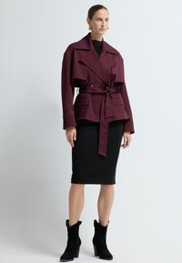 Burgunderrød skræddersyet jakke med bæltestrop i taljen, to forlommer, kombineret med en sort tætsiddende nederdel og sorte suedestøvler med hæl.