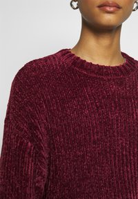 Vínový sametový svetr s žebrovanou texturou, kulatým výstřihem a pohodlným střihem. Jsou viditelné detaily na rukávech a límci.