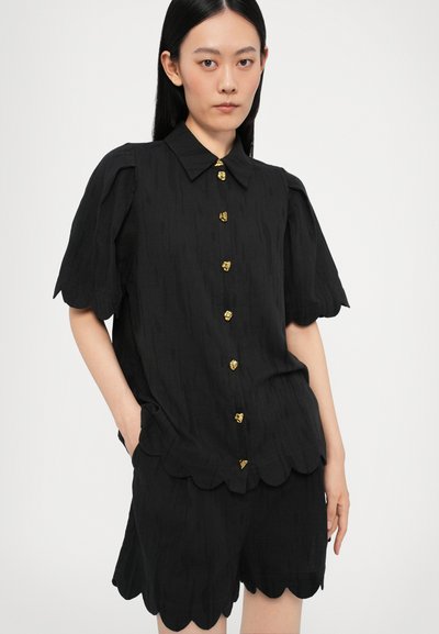 Schwarze Knopfbluse mit kurzen Puffärmeln und gewelltem Saum, versehen mit goldenem Herzknopf und strukturiertem Stoff. Kombiniert mit passenden Shorts.
