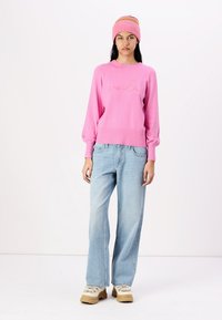 Pull en tricot fuchsia avec des manches bouffantes et un logo, associé à un jean large bleu clair et des bottes à plateforme beige chunky. Bonnet rayé.