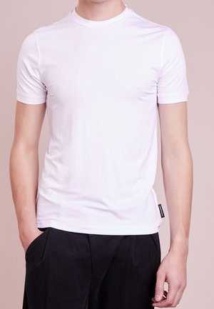 Mann trägt ein schlichtes weißes T-Shirt mit kurzen Ärmeln und schwarze Hose, eine Hand in der Tasche, neutraler beige Hintergrund.