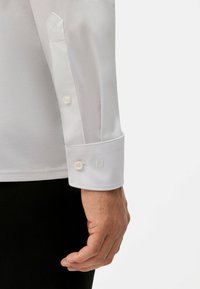 Chemise blanche à manches longues avec poignet pointu, fermeture à un bouton, broderie discrète sur le poignet, texture lisse et coupe ajustée.