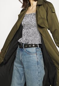 Trench-coat vert olive porté sur un haut smocké à carreaux noir et blanc, associé à un jean taille haute bleu clair et une ceinture noire avec des œillets métalliques.