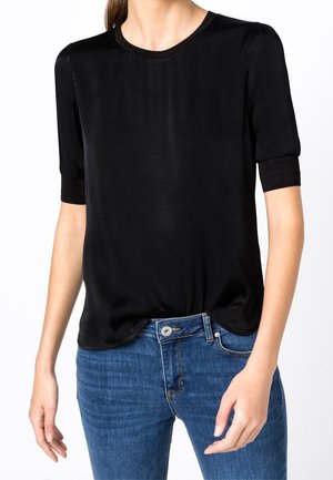 Femme portant un chemisier satiné noir à manches courtes, rentré dans un jean en denim bleu, vue des épaules aux hanches.