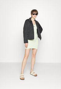 Veja corta preta sobre um vestido justo verde menta, combinado com sandálias bege. Modelo usando óculos escuros escuros, posando contra um fundo branco.