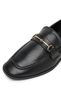 Czarne skórzane loafersy z płaskim noskiem, złotym detalem na wierzchu oraz subtelnymi przeszyciami wzdłuż krawędzi.