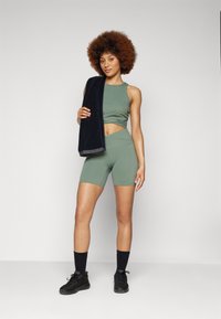Completo sportivo con un top corto aderente color oliva e pantaloni shorts a vita alta coordinati, abbinato a un asciugamano scuro appoggiato su una spalla.