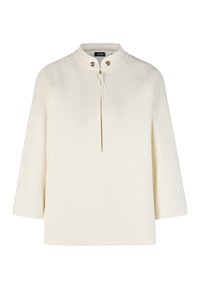 Blouse crème à manches longues avec petit col montant, fermeture éclair devant avec tirette, et deux boutons-pression métalliques près de l'encolure.
