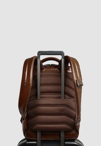 Mochila de cuero marrón con respaldo acolchado, correas ajustables y un mango retráctil. Presenta un tejido texturizado en la parte trasera y herrajes de metal.