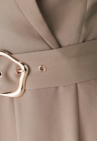 Bēša kleitas audums ar gludu faktūru, ar platu jostu, kas rotāta ar abstraktu zelta sprādzi un metāla acs detaļu.