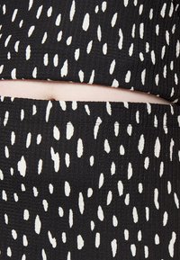 Tecido preto com uma superfície texturizada, apresentando um padrão de pontos brancos variados. O design inclui uma secção superior e inferior ligeiramente encurtada.