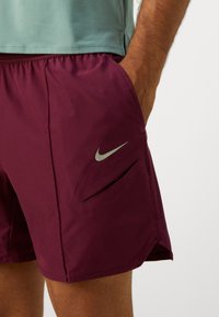 Bordeauxfarbene Sportshorts mit einem silbernen Nike-Logo, seitlichen Einschubtaschen und einem elastischen Bund, aus einem leichten, strukturierten Stoff.