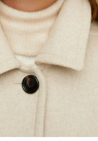 Abrigo de lana beige con un gran botón negro sobre un suéter de cuello alto de canalé color crema, mostrando el cuello y la parte superior del pecho.