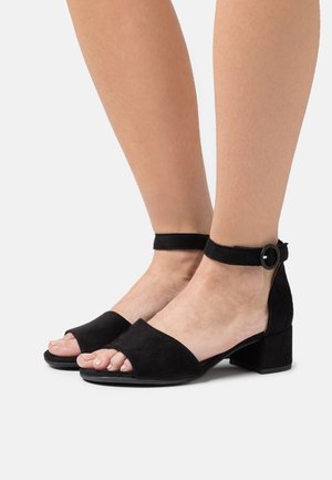 Sandales à talons en daim noir avec un design à bout ouvert, une sangle à la cheville et un talon bloc. Texture lisse avec un style minimaliste.