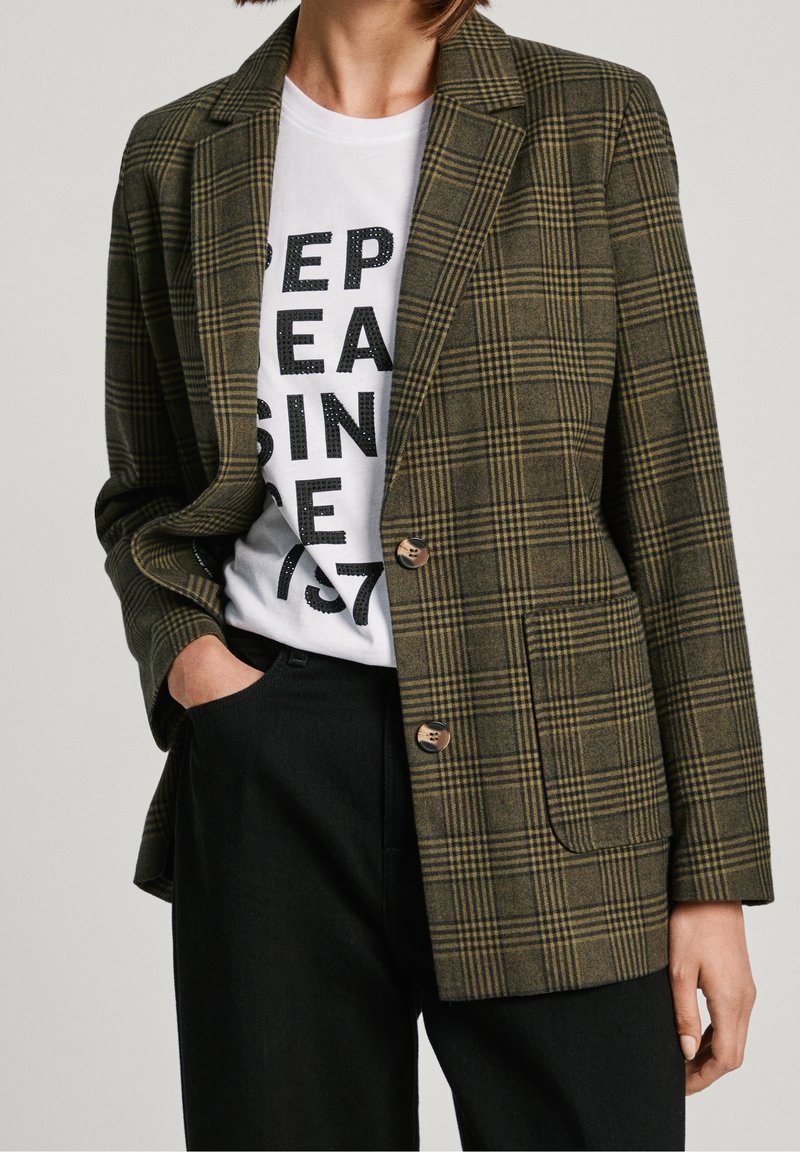 Person iført olivengrøn ternet blazer, hvidt grafisk t-shirt med sort tekst og sorte bukser, med den ene hånd i lommen.