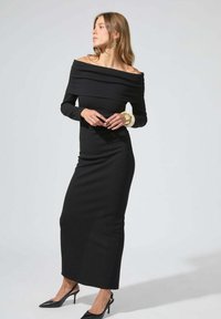 Robe noire décolletée épaules nues avec manches longues, ajustement près du corps et texture lisse. Associée à des talons pointus noirs et des accessoires dorés.