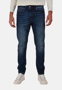 Jeans in denim di colore blu scuro con una vestibilità slim. Presentano dettagli cuciti, cinque tasche e una finitura leggermente sbiadita. Indossati con sneakers bianche.