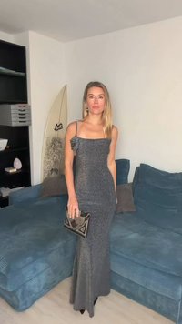 Γκρι, λαμπερό βραδινό φόρεμα με εφαρμοστή σιλουέτα, με διακοσμητικό λουλούδι στον ώμο. Κρατάει μεταλλική τσάντα φάκελο.