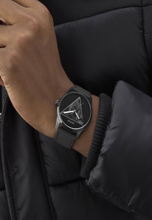 Schwarze analoge Uhr mit Gummiband, die ein dreieckig gemustertes Zifferblatt und silberne Akzente aufweist, getragen an einer Hand neben einer dunklen Jacke.