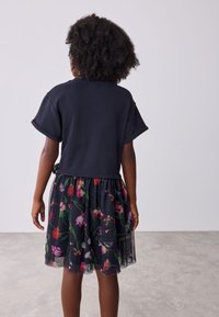 Schwarzes Kurzarmoberteil mit kurzem Schnitt, kombiniert mit einem mehrfarbigen Tüllrock mit Blumenmuster, transparentem Überwurf und elastischem Bund.