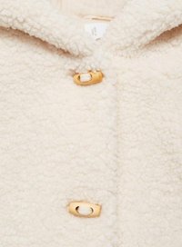 Pehmeä kermanvärinen takki, jossa on teksturoitu, pehmeä fleece-materiaali, hupullinen malli ja kultaiset varusteiden napit etuosassa.