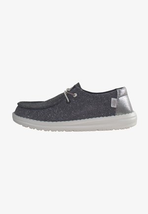 Zapatilla de lona gris con brillo, suela de goma blanca, puntera redonda y diseño de cordones, que cuenta con una pequeña etiqueta en el lateral.