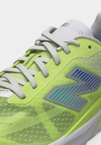 Κοντινή εικόνα ενός παπουτσιού τρεξίματος νέον κίτρινου και λευκού με λευκά κορδόνια και μπλε λογότυπο New Balance στο πλάι.