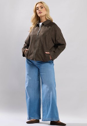 Lyshåret kvinde iført en mørkebrun jakke med lynlås, lyseblå vidde bukser og brune flade sko, stående med hænderne i lommerne.