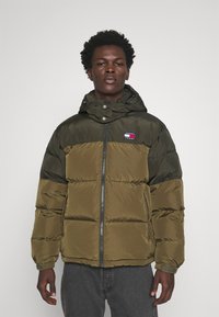 Chaqueta acolchada en verde oliva oscuro con cierre de cremallera en la parte delantera, capucha y puffs cosidos. Cuenta con un pequeño parche de logo en el pecho. Fabricada con material suave.