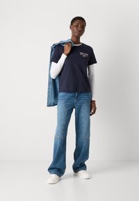 T-shirt in cotone blu navy con logo, sovrapposta a una camicia bianca a maniche lunghe. Jeans a gamba larga azzurri chiari con dettagli in patch. Sneakers bianche.