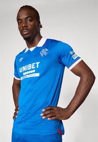 Blaues Fußballtrikot mit weißem Kragen, roten Akzenten und Sponsor Logos. Hergestellt aus einem glatten, leichten Stoff mit einer schmalen Passform.