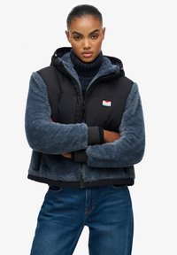 Gilet imbottito con corpo in nylon nero e maniche e cappuccio in pelliccia artificiale blu. Presenta una zip frontale e un patch con logo. Indossato sopra un dolcevita blu navy.