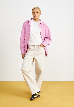 Pink corduroy shirt over a white t-shirt, paired with beige wide-leg pants and black shoes featuring yellow stripes. Set against a yellow background.