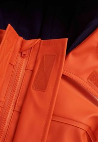 Tuva oranžā ūdensizturīgā jakā tuvplānā ar rāvējslēdzēju, Velcro lapiņām un teksturētu audumu. Iekšpusē ir tumša vilnas oderes.