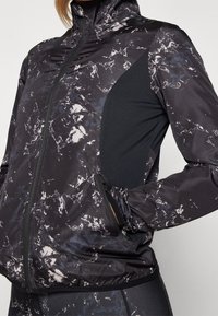 Černá sportovní bunda s mramorovým vzorem, vysokým límcem, zipovým uzávěrem, síťovanou panely na boku, elastickými manžetami a zipovými kapsami.