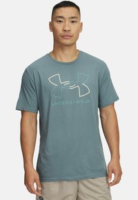 Teal kortærmet t-shirt med rund hals, prydet med et stort Under Armour-logo i lyse farver på forsiden. Blødt stof, afslappet pasform.