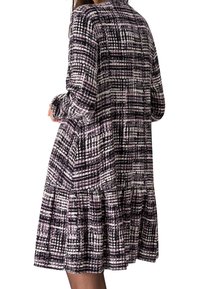 Femme portant une robe longueur genou à manches longues avec un motif à carreaux abstrait violet, noir et blanc, vue de dos.