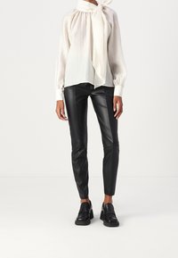 Blouse blanche plissée avec un col noué, associée à un pantalon en cuir noir ajusté et des mocassins épais noirs.