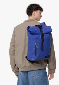 Rulltopprygsäck i livlig blå färg, med svarta remmar, en framfickan med dragkedja och ett slätt, hållbart tyg. Enkel, modern design.