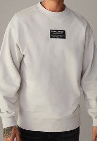 Sudadera blanca con cuello redondo, mangas largas y una etiqueta de parche negra que presenta el nombre de la marca y las coordenadas cosidas en la parte frontal.
