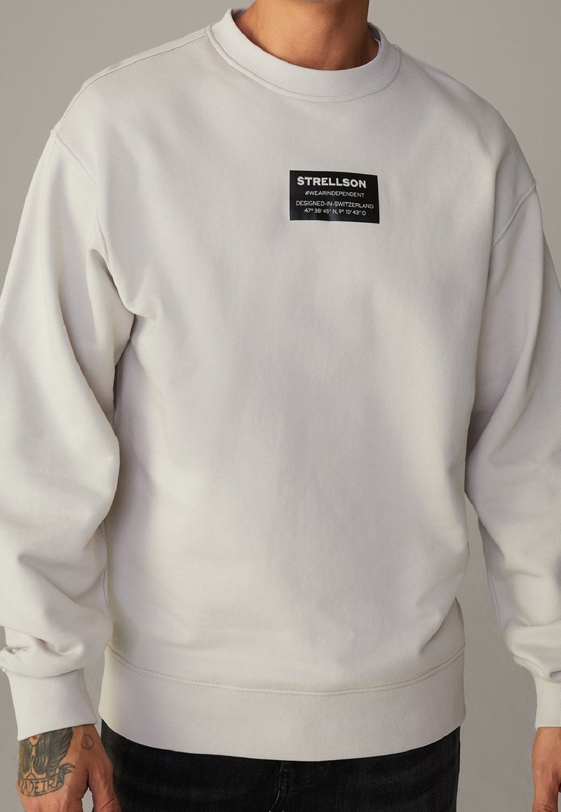 Sudadera blanca con cuello redondo, mangas largas y una etiqueta de parche negra que presenta el nombre de la marca y las coordenadas cosidas en la parte frontal.