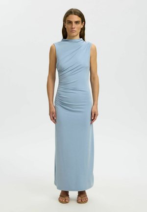 Femme debout dans une robe longue sans manches bleu clair avec fronces sur un côté, portant des sandales couleur beige, sur fond blanc uni.