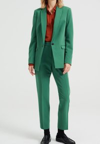 Zielony żakiet i dopasowane zwężane spodnie z gładkiej tkaniny. Pod spodem brązowa koszula z kołnierzykiem. Czarne mokasyny. Minimalistyczny design, bez wzorów.