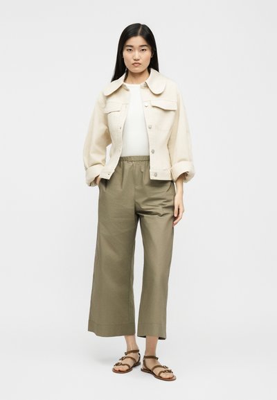 Γυναίκα που φοράει ένα κρεμ cropped σακάκι, λευκό τοπ, ελιάς φαρδιά cropped παντελόνια και καφέ σανδάλια με λουράκια, στέκεται μπροστά σε ένα απλό φόντο.