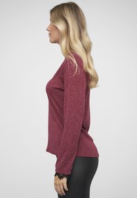 Bordeauxrotes Langarmshirt in Marl-Optik mit leicht geschwungenem Saum und Spitzenakzenten an den Manschetten, kombiniert mit schwarzen, enganliegenden Hosen.