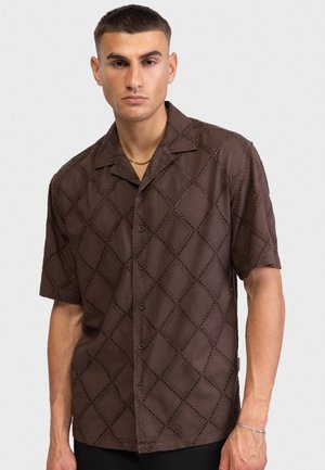 Mężczyzna ubrany w ciemnobrązową, krótkorękawową koszulę zapinaną na guziki z wzorem otworów w kształcie diamentów oraz złoty łańcuszek na gładkim tle.