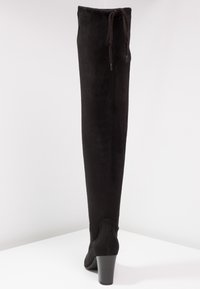 Botte noire montante au-dessus du genou en daim, dotée d'un talon haut, d'une surface légèrement texturée et d'un détail noué en haut.