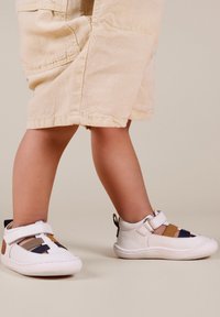 Zapatos blancos para niños con rayas multicolores en azul, marrón y beige; con una tira de Velcro y punta redondeada.