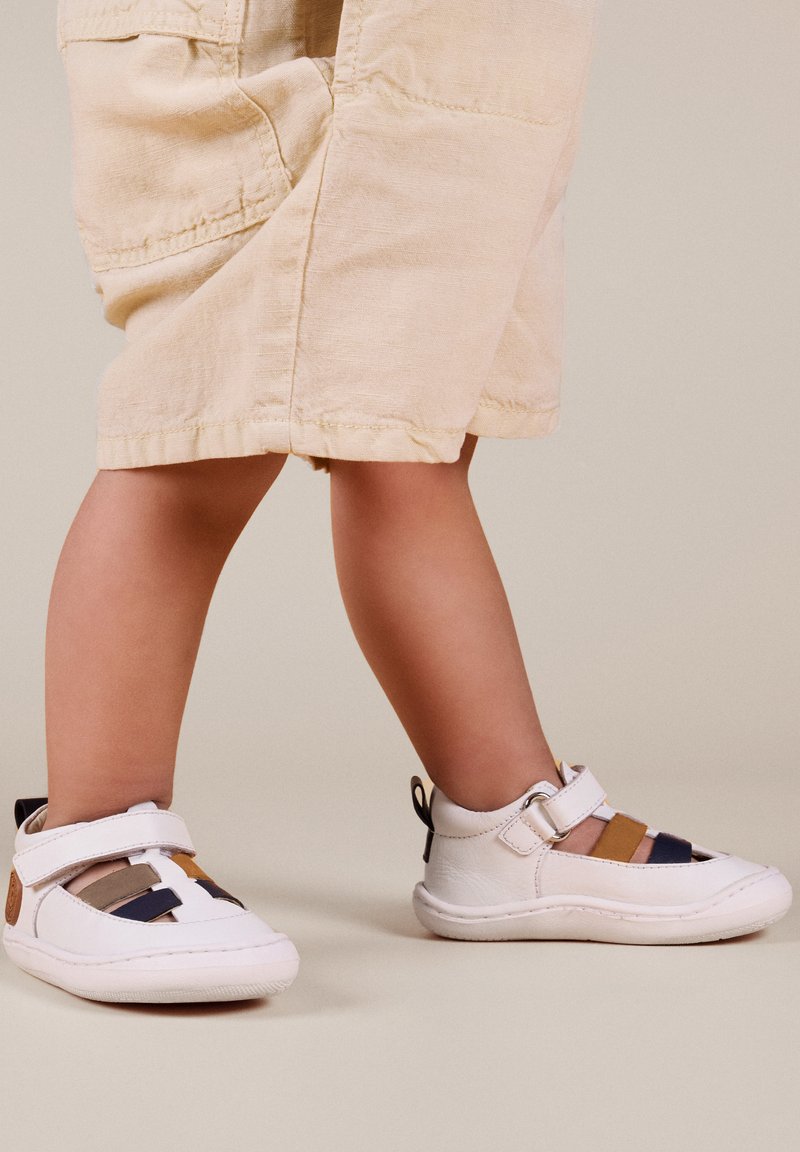 Zapatos blancos para niños con rayas multicolores en azul, marrón y beige; con una tira de Velcro y punta redondeada.